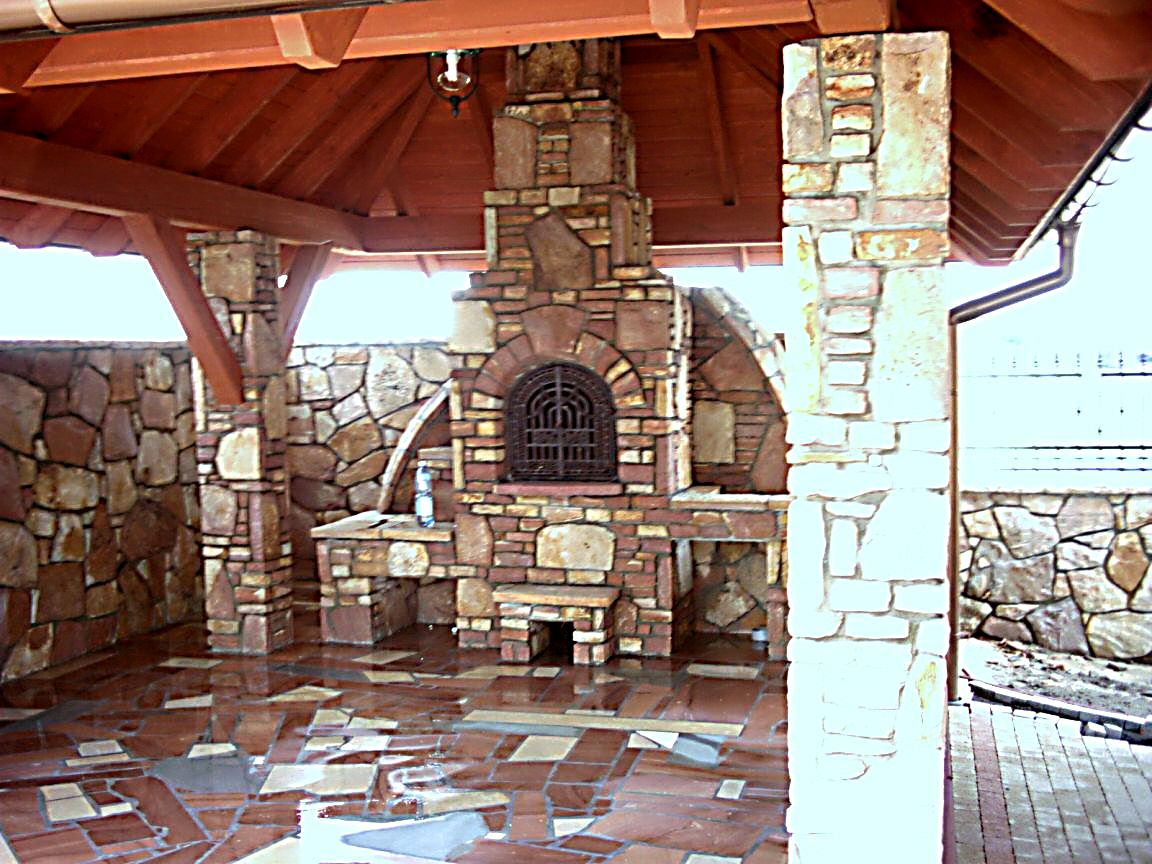 Kamienna konstrukcja z paleniskiem i grillem w zadaszonej przestrzeni, wykonana z nieregularnych kamieni w odcieniach brązu i beżu, z dekoracyjną kratą i błyszczącą podłogą.