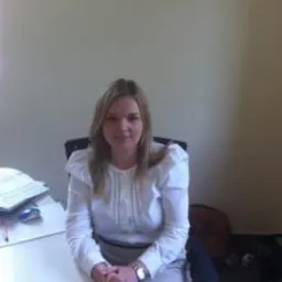 Kobieta w białej bluzce siedzi za biurkiem z dokumentami i ołówkami, z rękami złożonymi na blacie. Proste tło w jasnym kolorze.