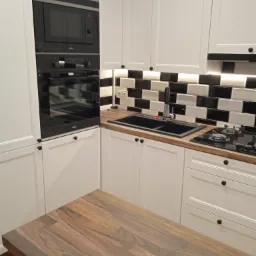 Nowoczesna biała kuchnia z czarnymi akcentami: zabudowa z piekarnikiem i mikrofalówką Amica, czarny zlew i bateria, płyta gazowa, blat drewniany z widocznym usłojeniem, czarno-białe płytki nad blatem.