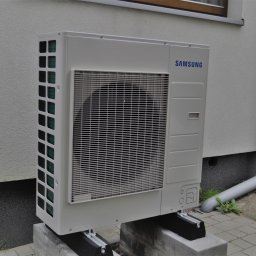 ZENE.pl - Zewnętrzna jednostka klimatyzacji Samsung zamontowana na betonowych bloczkach przy ścianie budynku z rynną spustową.