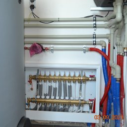 ZENE.pl - Rozdzielacz podłogowy z zaworami, rurami w izolacji czerwonej i niebieskiej, oraz rurami białymi, zamontowany na ścianie w pomieszczeniu technicznym. Widoczny fragment szarego zbiornika. Na ścianie...