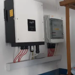 Biała obudowa falownika FOX ESS z czarnym wyświetlaczem i podłączonymi przewodami elektrycznymi oraz skrzynka zabezpieczeń na ścianie wewnątrz pomieszczenia.