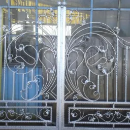 Metalowa brama z dekoracyjnymi, kutymi elementami w kształcie liści i zawijasów, na tle okna z szybami. Szare wykończenie, symetryczny wzór.