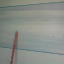 Ściana łazienki z ułożonymi, poziomymi płytkami w odcieniach bieli i błękitu, widoczna poziomica i zabezpieczone folią miejsce na gniazdko elektryczne.