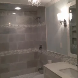 Wykończona łazienka w szarościach: płytki imitujące kamień, dekoracyjne pasy mozaiki, biała umywalka z szafką i lustro z półkami. Oświetlenie punktowe i kinkiet.