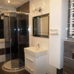 Nowoczesna łazienka z kabiną prysznicową z chromowanymi elementami, umywalką z szafką, grzejnikiem drabinkowym i lustrem w czarnej ramie, wykończona płytkami w odcieniach bieli i szarości,...