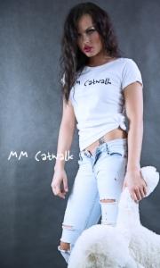 Kobieta w białej koszulce z napisem 'I'm Catwalk' i podartych jeansach, pozująca z pluszowym misiem na szarym tle. Widoczny tatuaż na brzuchu i logo 'MM Catwalk'.