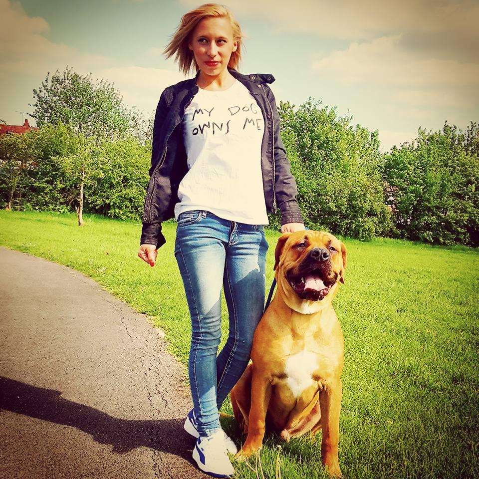 Blondynka w jeansach i kurtce pozuje z psem na trawie. Na koszulce napis 'My dog owns me'. Słoneczny dzień w parku.