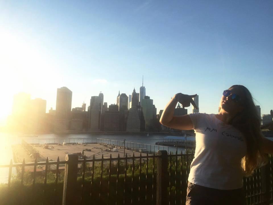 Kobieta w okularach przeciwsłonecznych, w koszulce z nadrukiem 'MM Catwalk', pozuje na tle panoramy Manhattanu w słoneczny dzień.