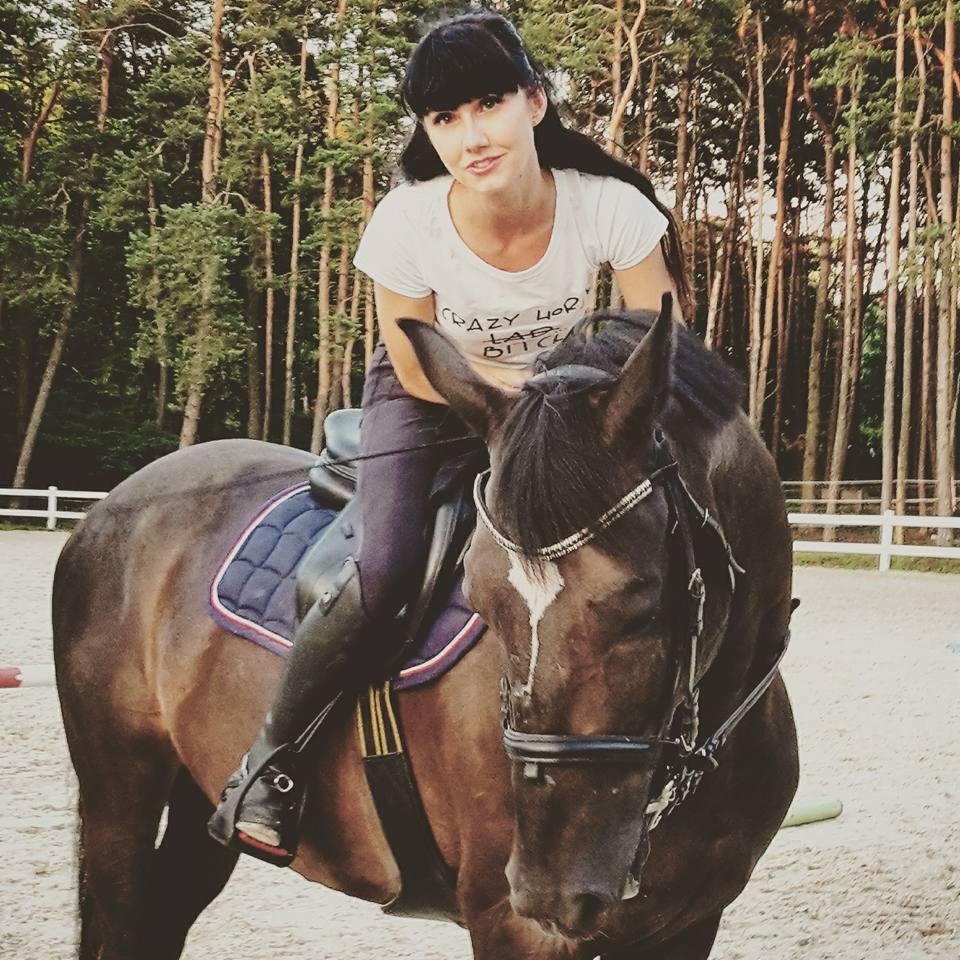 Kobieta w białej koszulce z nadrukiem siedzi na brązowym koniu na piaszczystym padoku z lasem w tle. Nadruk na koszulce jest częściowo widoczny.