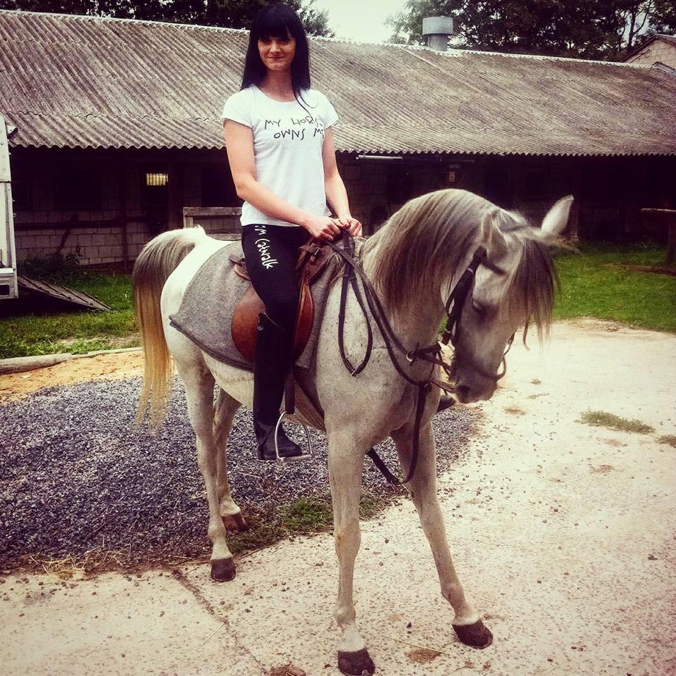 Kobieta na koniu w koszulce z nadrukiem 'My horse owns me'. Nadruk na odzieży w plenerze, stajnia w tle. Ujęcie z bliska, naturalne światło.