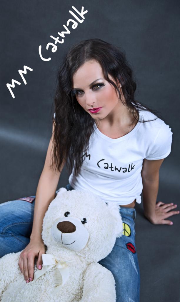 Kobieta w białej koszulce z nadrukiem 'mm Catwalk' i jeansach z naszywkami, siedząca z pluszowym misiem na szarym tle.