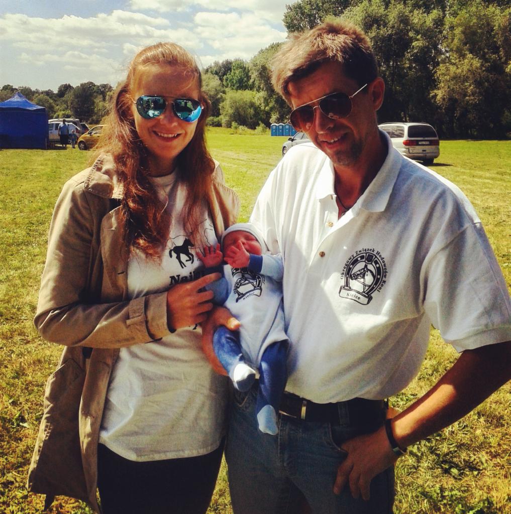 Rodzina prezentuje koszulki z nadrukiem: mama w t-shircie z koniem, tata w polo z logo, dziecko w body z grafiką. Plener, słoneczny dzień.