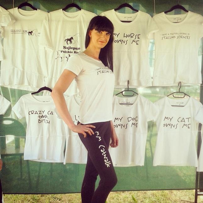 Kobieta prezentuje białą koszulkę z napisem na tle ekspozycji koszulek z różnymi nadrukami, w tym 'My horse owns me' i 'Crazy cat lady'.