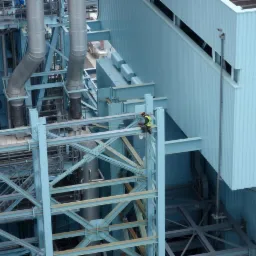 Pracownik w kasku i kamizelce na wysokości, wykonuje prace na metalowej konstrukcji przy budynku z blachy trapezowej w kolorze błękitnym.