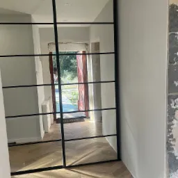 Przesuwne lustrzane drzwi z czarnymi szprosami, widok na korytarz z drewnianą podłogą i otwartymi drzwiami wejściowymi, ściana w trakcie wykończenia.