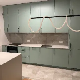 Nowoczesna kuchnia z frontami w kolorze miętowym, czarnymi uchwytami i blatem w odcieniach szarości, oświetlona designerską lampą z białymi elementami.