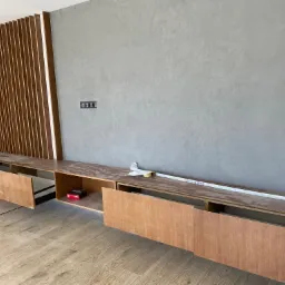 Wnętrze w trakcie montażu: Podwieszana szafka RTV z drewnianymi frontami i dekoracyjnymi pionowymi listwami na ścianie z szarą fakturą, widoczne narzędzia i materiały budowlane.