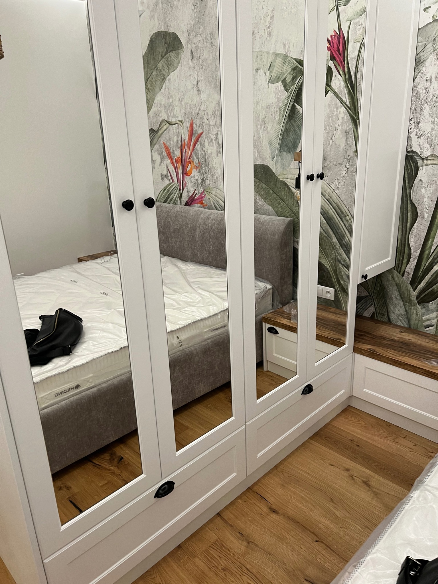 Biała szafa wnękowa z lustrami, odbijająca łóżko i tapetę w tropikalne liście, z drewnianym blatem i szafkami po prawej stronie.