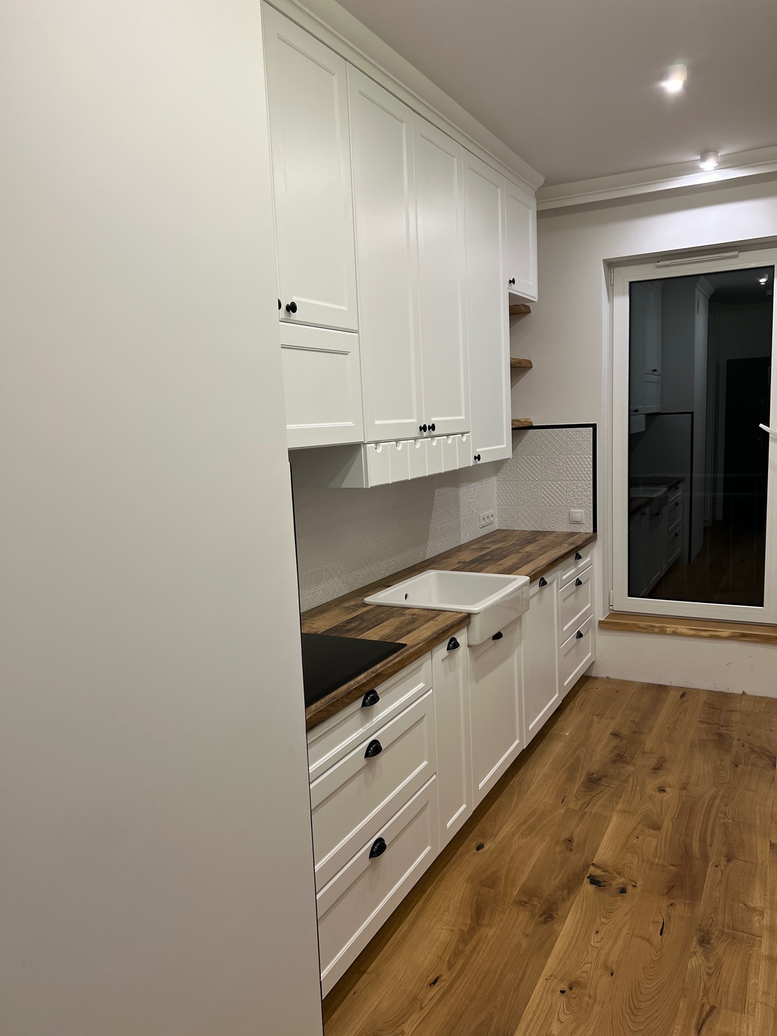 Jasna kuchnia z białymi szafkami, drewnianym blatem i podłogą, widok z boku na umywalkę i okno.