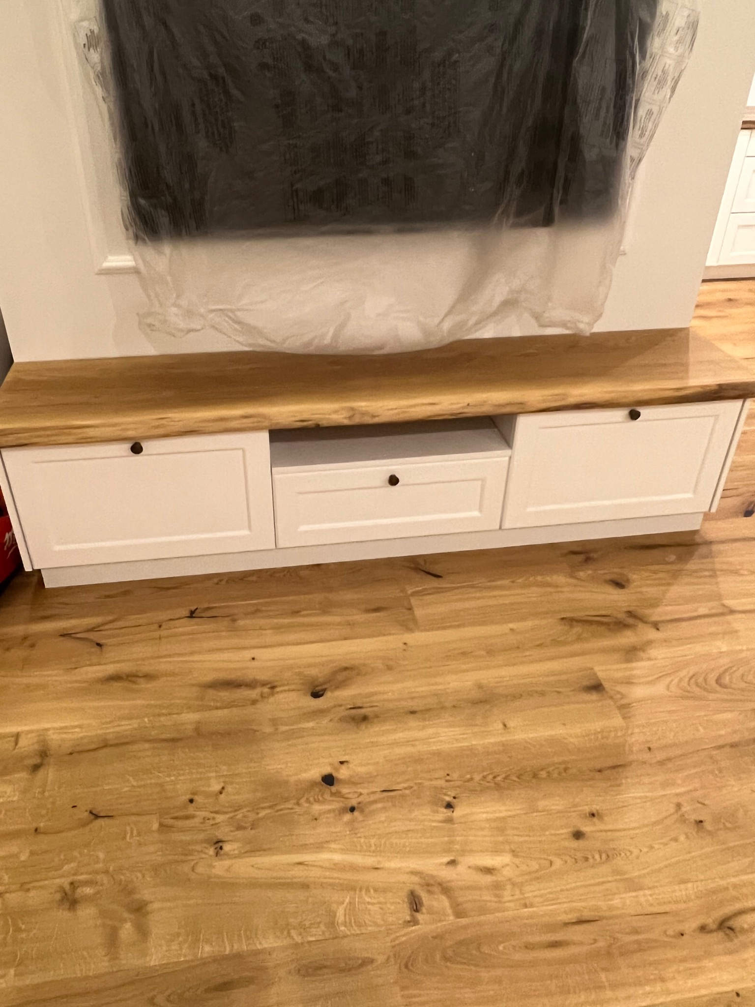 Szafka RTV z białymi frontami i blatem z litego drewna dębowego, wykonana na wymiar w Radomsku, z widoczną folią ochronną na ścianie.