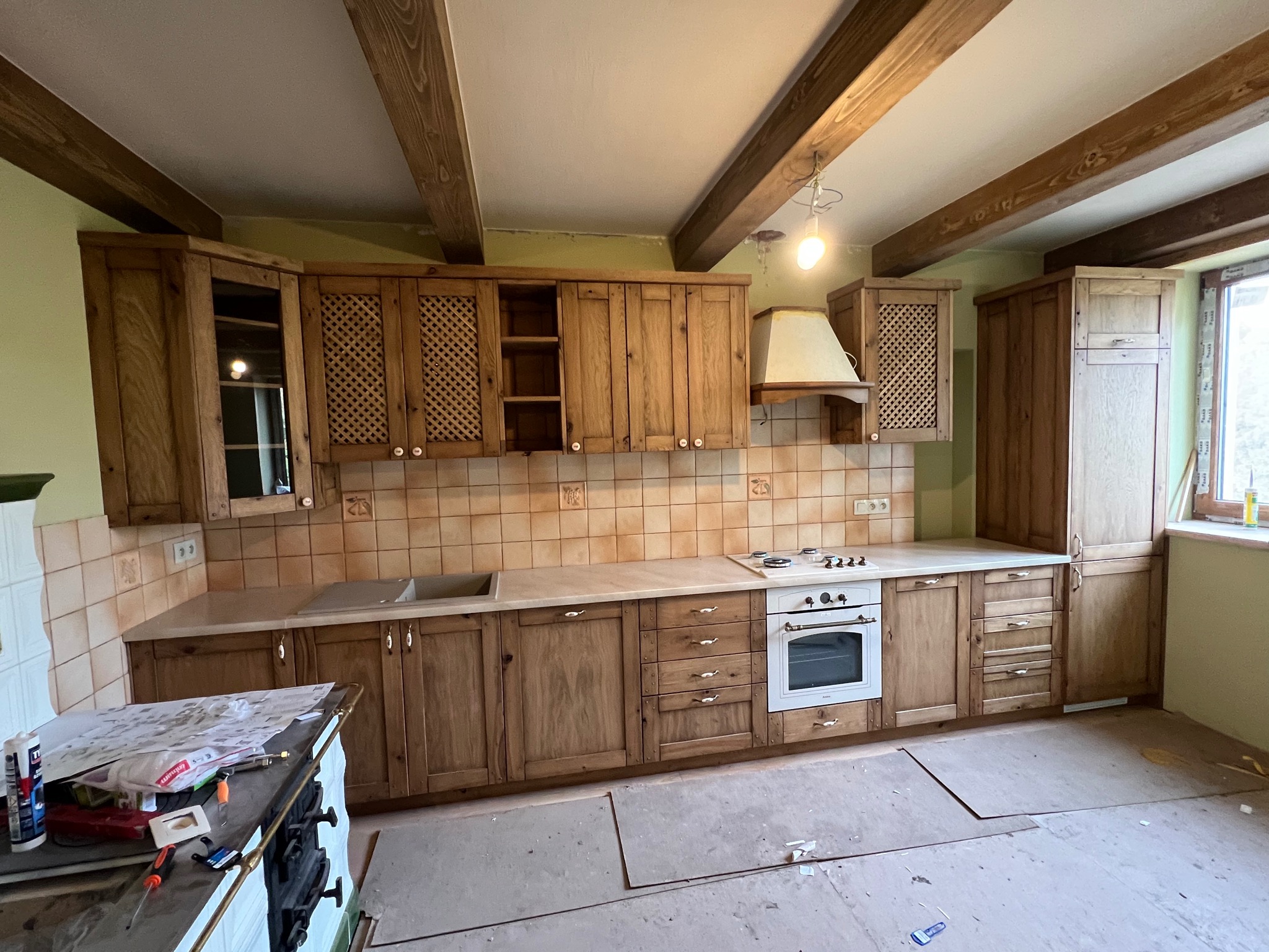Rustykalna kuchnia z drewnianymi szafkami i beżowymi płytkami, widoczne drewniane belki stropowe i rozłożone narzędzia na blacie, sugerujące etap montażu.