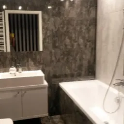 Nowoczesna łazienka z szarymi płytkami imitującymi marmur, białą wanną i umywalką z szafką. W lustrze odbija się grzejnik drabinkowy. Oświetlenie sufitowe.