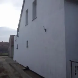 Świeżo otynkowana, biała ściana budynku z małymi oknami, widoczna z perspektywy bocznej. W tle fragment dachu i sąsiedni budynek. Pochmurne niebo.