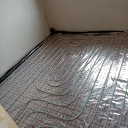 Białe rury ogrzewania podłogowego ułożone na folii z czerwoną kratką, widoczne czarne rury brzegowe i fragment okna dachowego w pokoju na poddaszu.