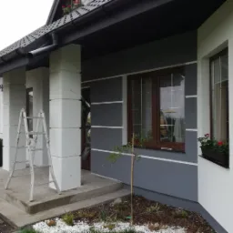 Wykończony fragment elewacji domu z szarą ścianą, białymi narożnikami i brązowymi oknami, z drabiny opartą o kolumnę werandy i osobą w tle.