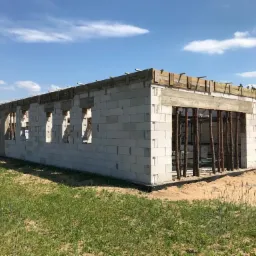 Wznoszenie ścian z betonu komórkowego Ytong z widocznym zbrojeniem nadproży okiennych i deskowaniem stropu garażu, w tle zabudowa jednorodzinna i błękitne niebo.