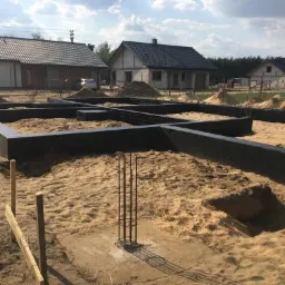 Przygotowany fundament pod dom z izolacją przeciwwilgociową, widoczne zbrojenia, w tle budynki w różnym stadium budowy.