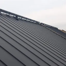 Nowe, grafitowe pokrycie dachu z blachy na domu jednorodzinnym, widok z rusztowania podczas montażu, z zamontowanymi barierkami śniegowymi.