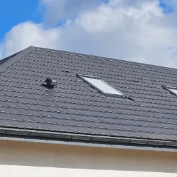 Szary dach z dachówki ceramicznej z dwoma oknami dachowymi, kominem i rynną, na tle błękitnego nieba z chmurami.