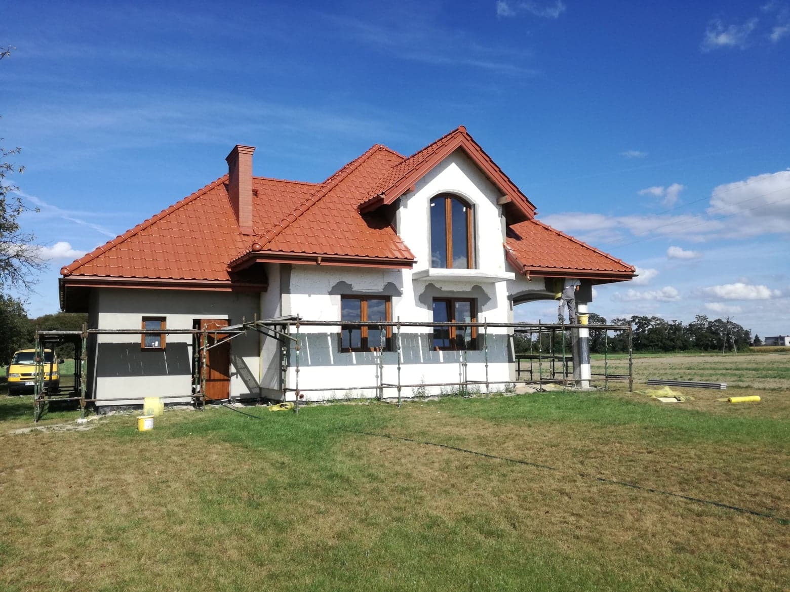 Dom murowany w trakcie budowy, z czerwoną dachówką, otynkowany na biało, z rusztowaniami i widocznym fragmentem pracownika na rusztowaniu, na tle błękitnego nieba i zielonego trawnika.
