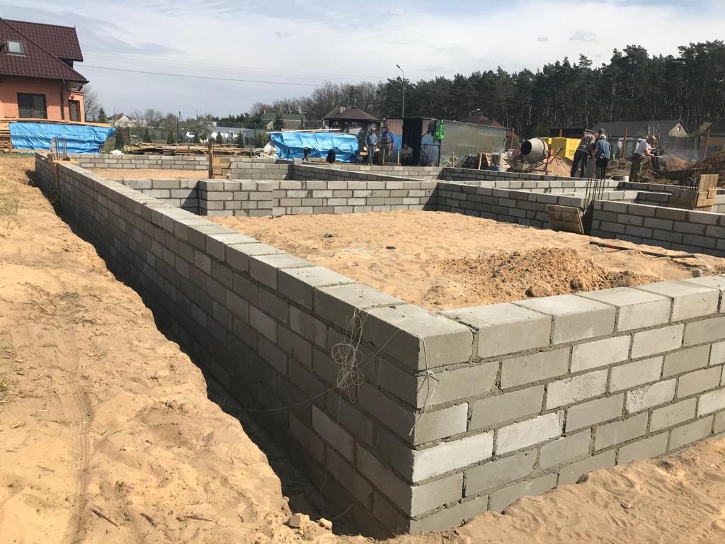 Wznoszenie fundamentów domu z bloczków betonowych; widoczne zbrojenia pionowe, piasek wewnątrz obrysu fundamentów, pracownicy budowlani w tle.