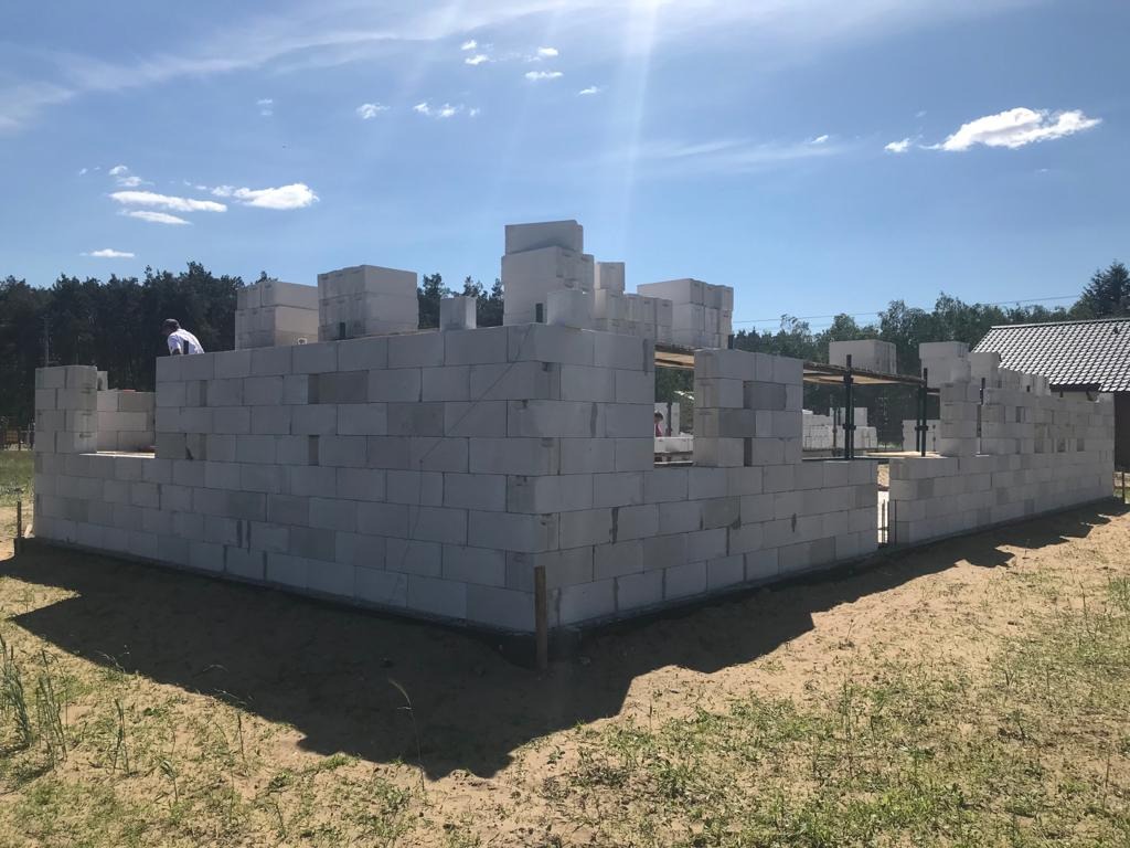 Wznoszone ściany domu z bloczków betonowych na piaszczystym terenie, widoczny pracownik w białej koszulce, w tle las i fragment dachu sąsiedniego budynku.