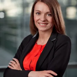 Uśmiechnięta kobieta w czarnej marynarce i czerwonej bluzce, z założonymi rękami na tle szklanej ściany biurowca.