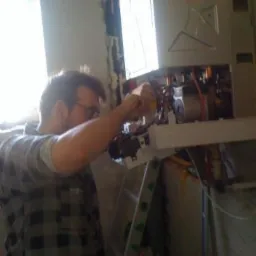 Mężczyzna w koszuli w kratę montuje piec gazowy przy oknie, stojąc na drabinie. Widoczne narzędzia i fragment ściany w trakcie remontu.