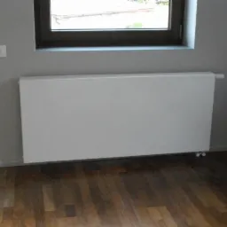 Biały, płaski grzejnik panelowy zamontowany na szarej ścianie z oknem i gniazdami elektrycznymi, na tle parkietu. Widok z niskiego kąta.