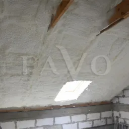 Poddasze po natrysku izolacji, widoczne drewniane belki stropowe i okno dachowe, ściany z białego bloczka betonowego.