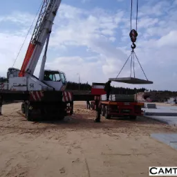 Żuraw samochodowy w trakcie podnoszenia betonowej płyty na przyczepę, widoczne podpory żurawia, piaskowe podłoże i ułożone płyty w tle.