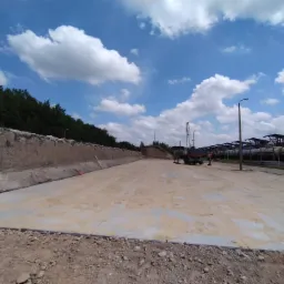 Przygotowany teren pod budowę z widocznymi betonowymi ścianami oporowymi, częściowo utwardzoną nawierzchnią i ciężkim sprzętem budowlanym w tle, pod błękitnym niebem z chmurami.