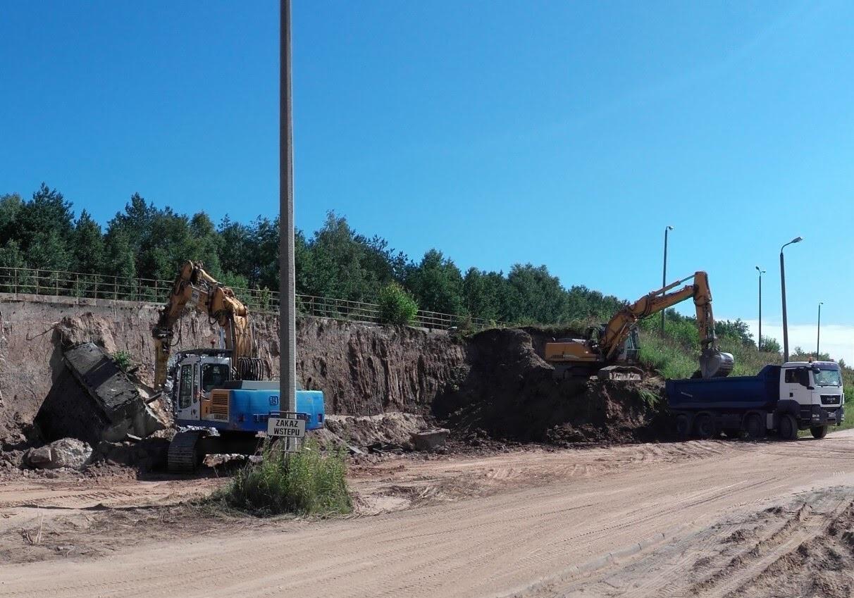 Żółto-niebieska koparka Liebherr kruszy betonowy element przy wykopie, w tle druga koparka i ciężarówka MAN, tabliczka 'Zakaz Wstępu', teren budowy.