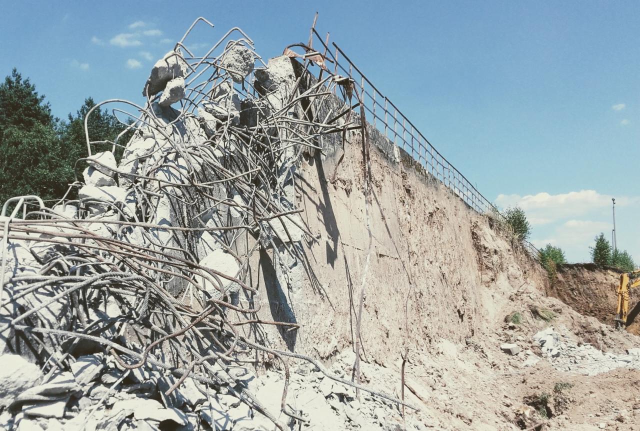 Zniszczony betonowy mur oporowy z wystającą stalową armaturą, widoczny wykop w ziemi i fragment maszyny budowlanej w tle.