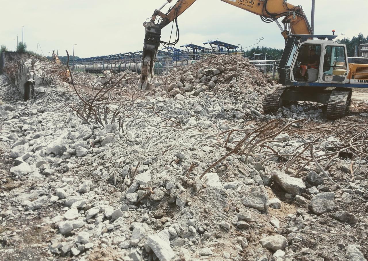 Żółta koparka hydrauliczna kruszy betonowy mur młotem pneumatycznym, w tle widoczne fragmenty konstrukcji stalowej oraz gruzowisko z wystającymi prętami zbrojeniowymi.