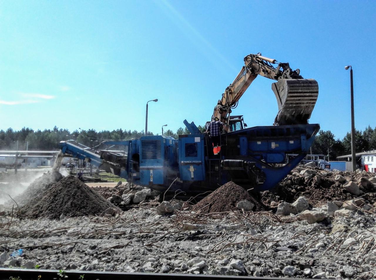 Niebieska kruszarka mobilna Terex Pegson w trakcie pracy na placu budowy, przetwarzająca gruz na frakcje, z widocznym operatorem w kabinie i stertami materiału na tle lasu.