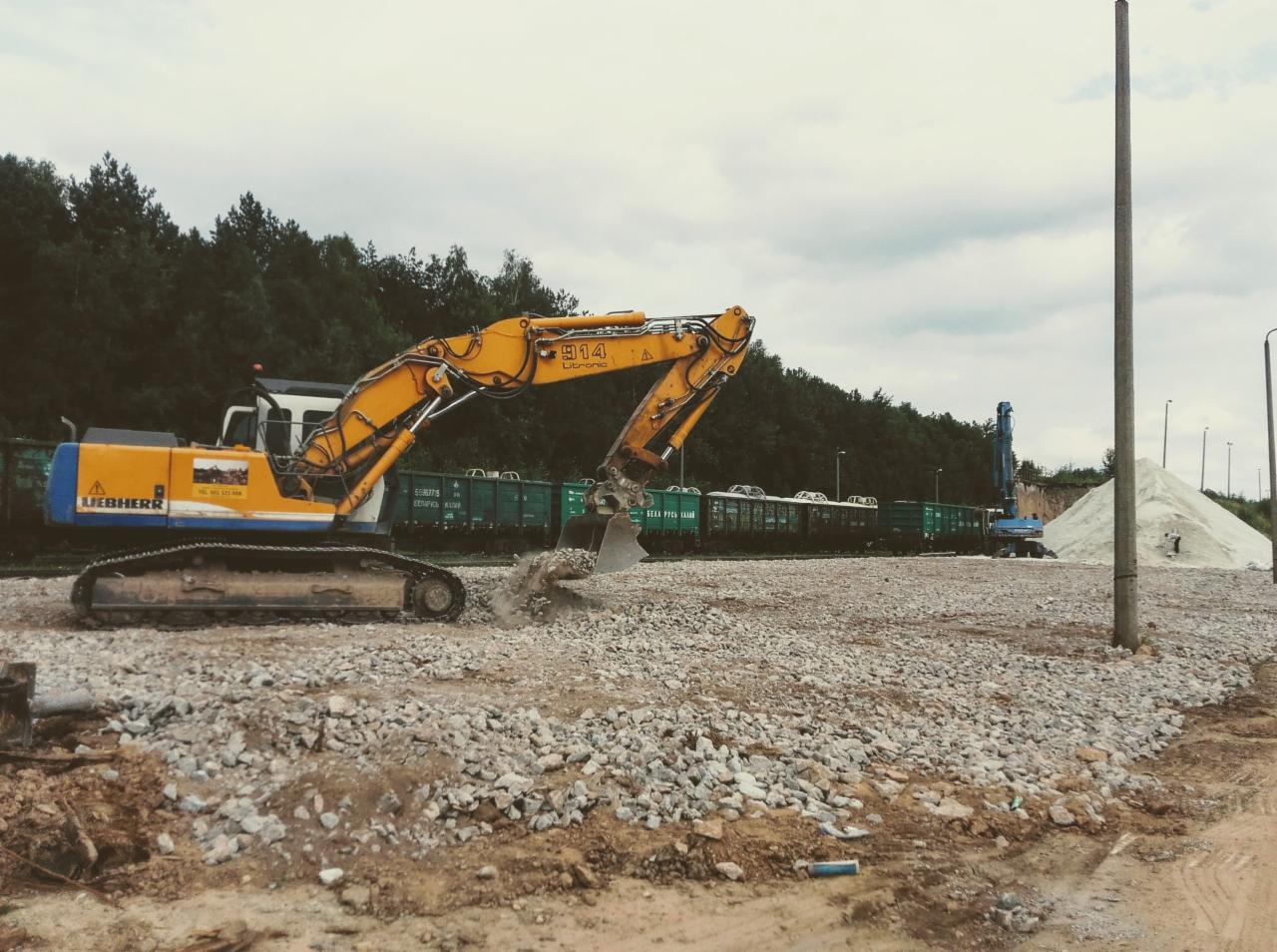 Żółto-niebieska koparka Liebherr 914 Litronic na placu budowy, przesypuje materiał na utwardzoną nawierzchnię z gruzu, w tle zielone wagony kolejowe i hałda piasku.