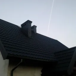 Nowy, ciemny dach z blachodachówki trapezowej i dwoma kominami z cegły na tle jasnego nieba z śladem kondensacyjnym samolotu.