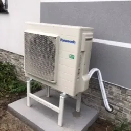 Zewnętrzna jednostka klimatyzacyjna Panasonic zamontowana na metalowym stelażu, ustawiona na betonowej podstawie przy szarej ścianie budynku z poziomymi pasami.